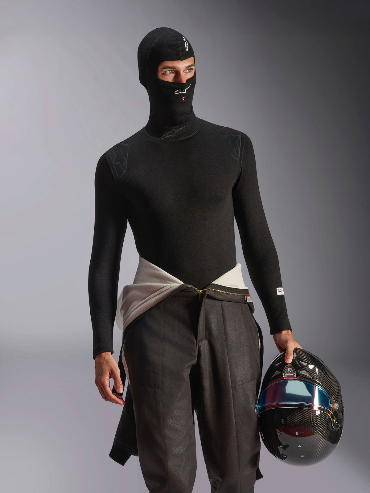Modelo masculino luciendo el pasamontañas Alpinestars ZX Evo V3 en negro, con logotipos blancos en la frente y el mentón, combinado con una prenda interior de manga larga negra y sujetando un casco de carreras de fibra de carbono, equipamiento profesional ignífugo para automovilismo
