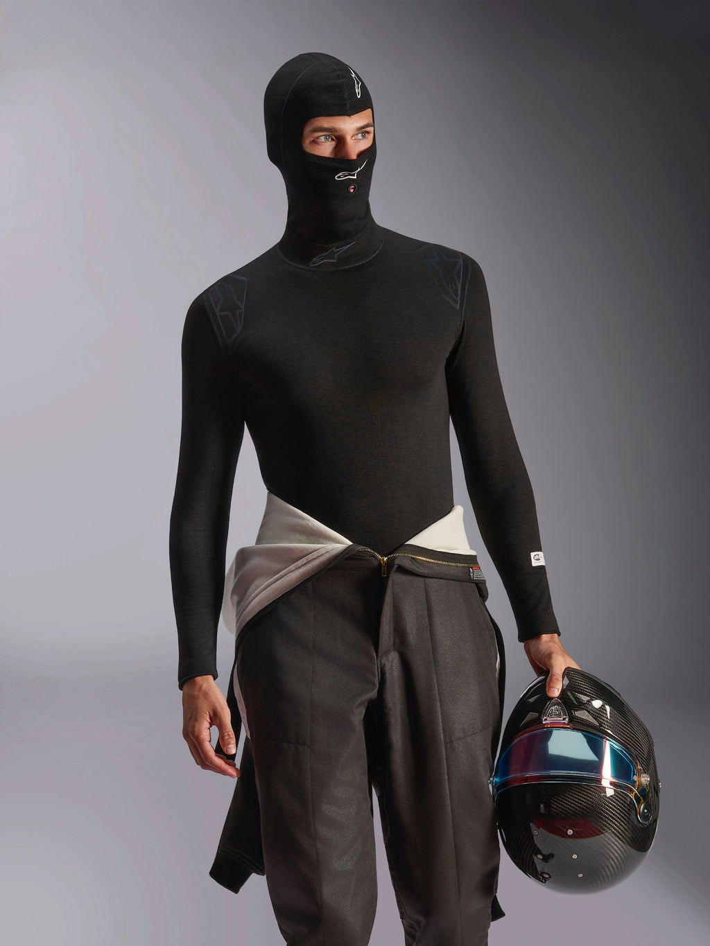 Modelo masculino luciendo el pasamontañas Alpinestars ZX Evo V3 en negro, con logotipos blancos en la frente y el mentón, combinado con una prenda interior de manga larga negra y sujetando un casco de carreras de fibra de carbono, equipamiento profesional ignífugo para automovilismo