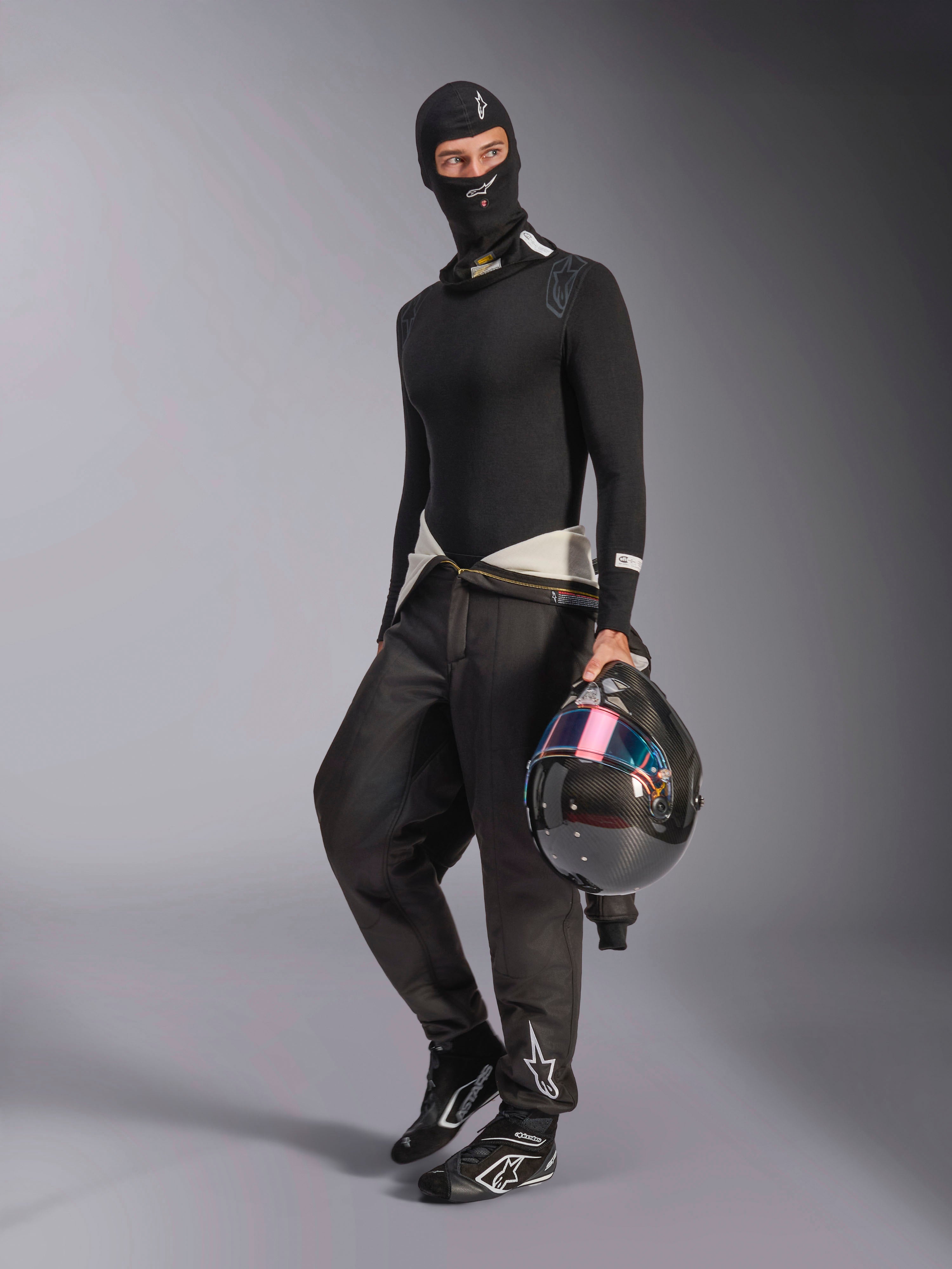 Pasamontañas Alpinestars ZX Evo V3, negro, usado por un piloto con equipo completo de capas base para automovilismo, con el logo blanco en la frente, sosteniendo un casco de fibra de carbono con visera iridiscente, ropa profesional de automovilismo resistente al fuego.