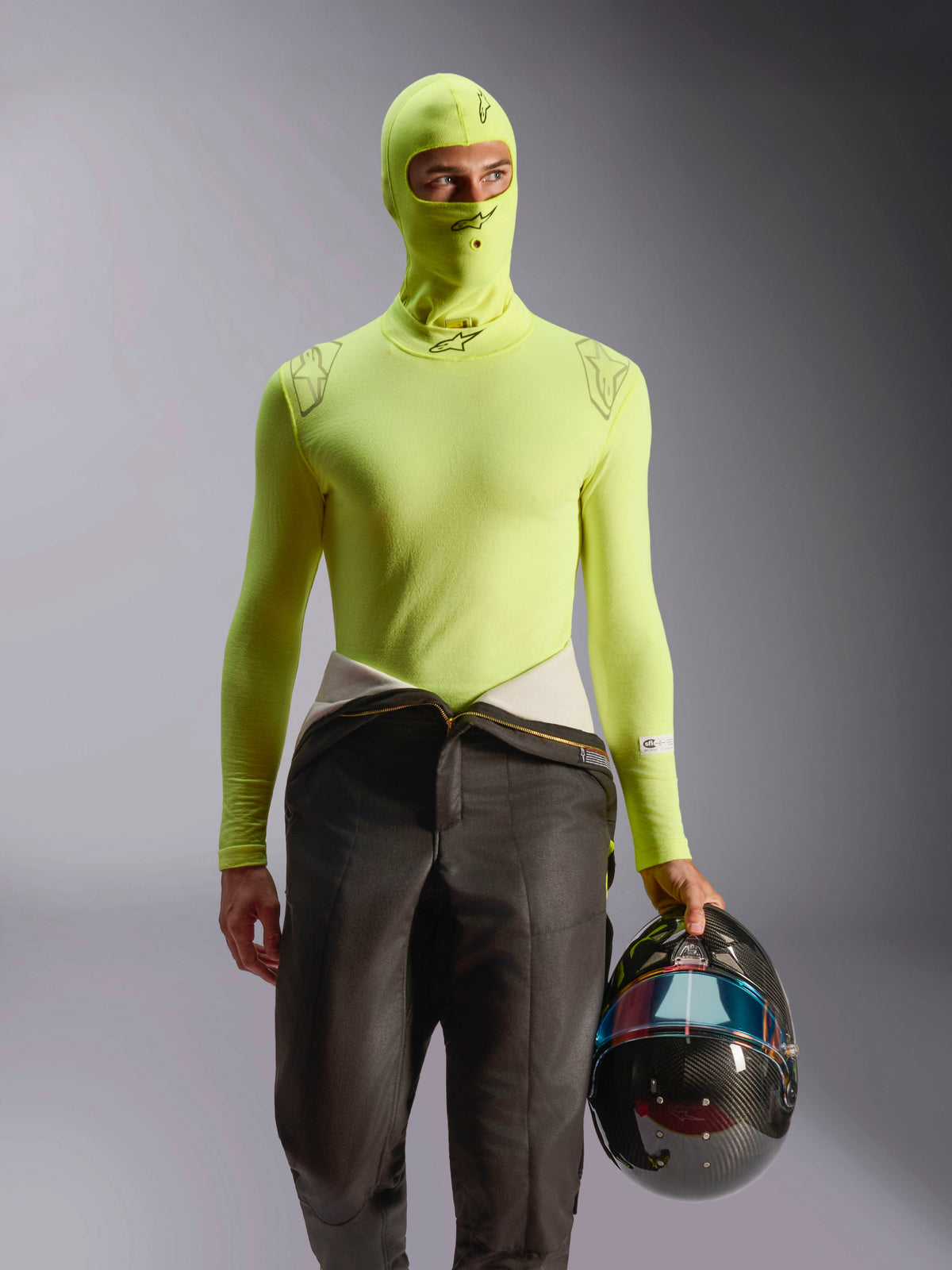 Pasamontañas Alpinestars ZX Evo V3, capa base para automovilismo, amarillo fluo, combinado con camiseta de manga larga y pantalones de traje de carreras, modelo sujetando un casco de fibra de carbono con visera iridiscente, equipo profesional de automovilismo resistente al fuego