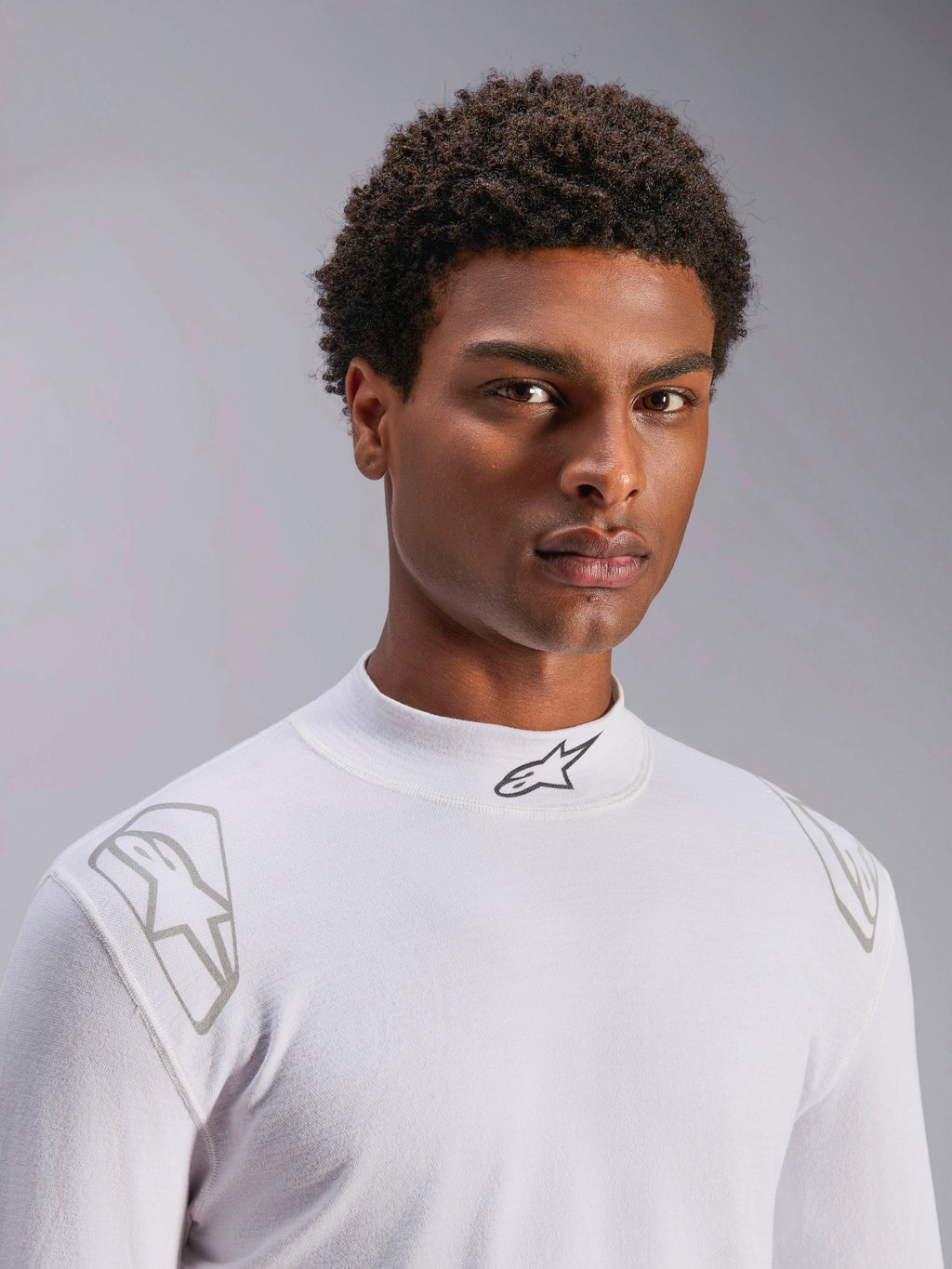 Alpinestars ZX Evo V3 Top - Manga Larga, capa base de automovilismo blanca, usada por un modelo masculino, cuello alto con logo Alpinestars negro, gráficos plateados en los hombros, ropa interior técnica ligera y resistente a las llamas para deportes de motor