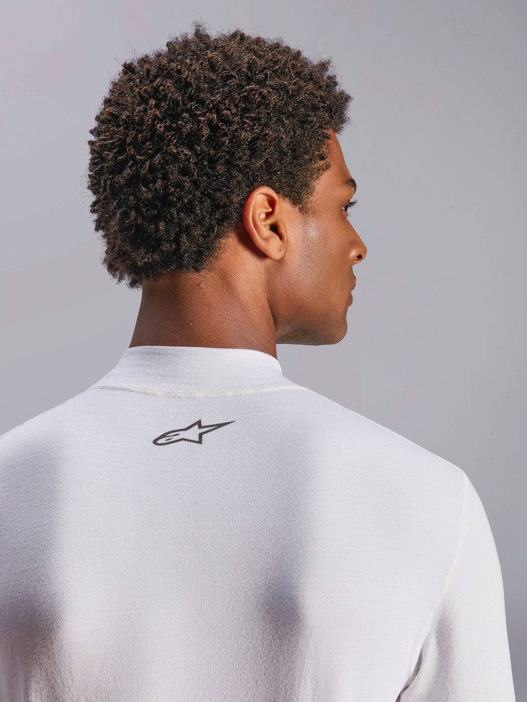 Alpinestars ZX Evo V3 Top, capa base de manga larga para automovilismo, blanco, primer plano de la zona posterior del cuello con el logotipo negro de Alpinestars, tejido técnico resistente a las llamas, usado por un modelo con cabello oscuro y rizado