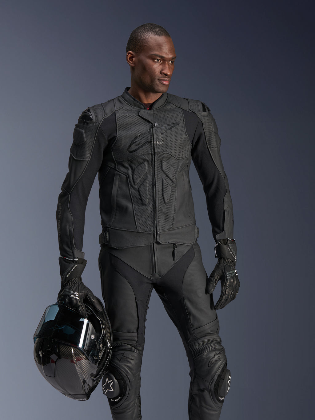 Chaqueta de cuero Alpinestars Dusk Airflow, negra, usada por un piloto masculino con pantalones de cuero y guantes a juego, sosteniendo un casco negro, con paneles de cuero perforado y protección integrada para conducción deportiva en moto.
