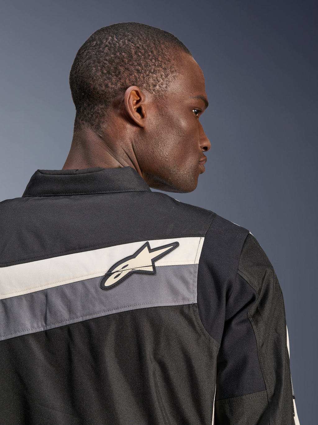Chaqueta Alpinestars T-Dyno WR, chaqueta textil para moto, negro, gris oscuro y crudo, primer plano de la parte superior de la espalda y el hombro con franjas horizontales y logotipo de la estrella de Alpinestars, usada por un modelo, diseño de inspiración heritage para conducción en carretera