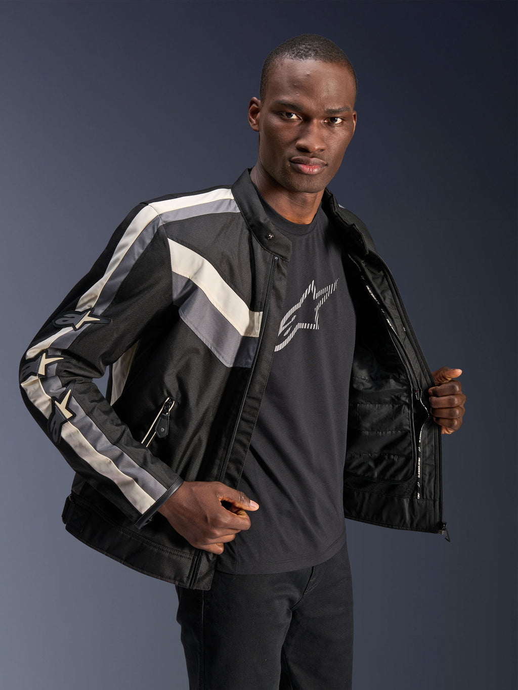 Chaqueta Alpinestars T-Dyno WR, negro, gris oscuro y crudo, chaqueta textil de moto con ajuste de motociclista de inspiración vintage, detalles de rayas en el pecho y las mangas, usada sobre una camiseta negra con gráfico, equipamiento para conducción urbana