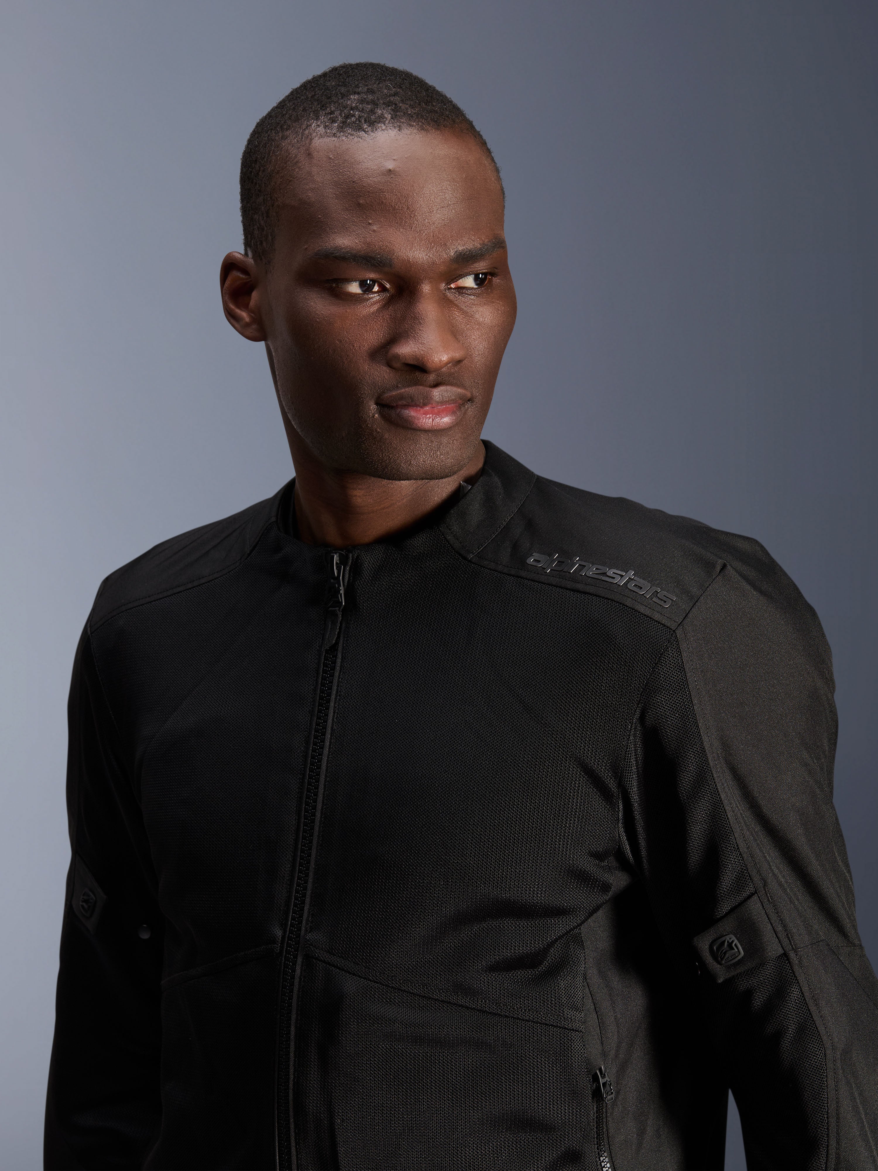 Chaqueta Alpinestars C-1 Air, negra, chaqueta de moto urbana con paneles de malla técnica, usada por un modelo, con un sutil logotipo de Alpinestars de transferencia de calor en el hombro, diseñada para el flujo de aire y comodidad ligera en climas cálidos