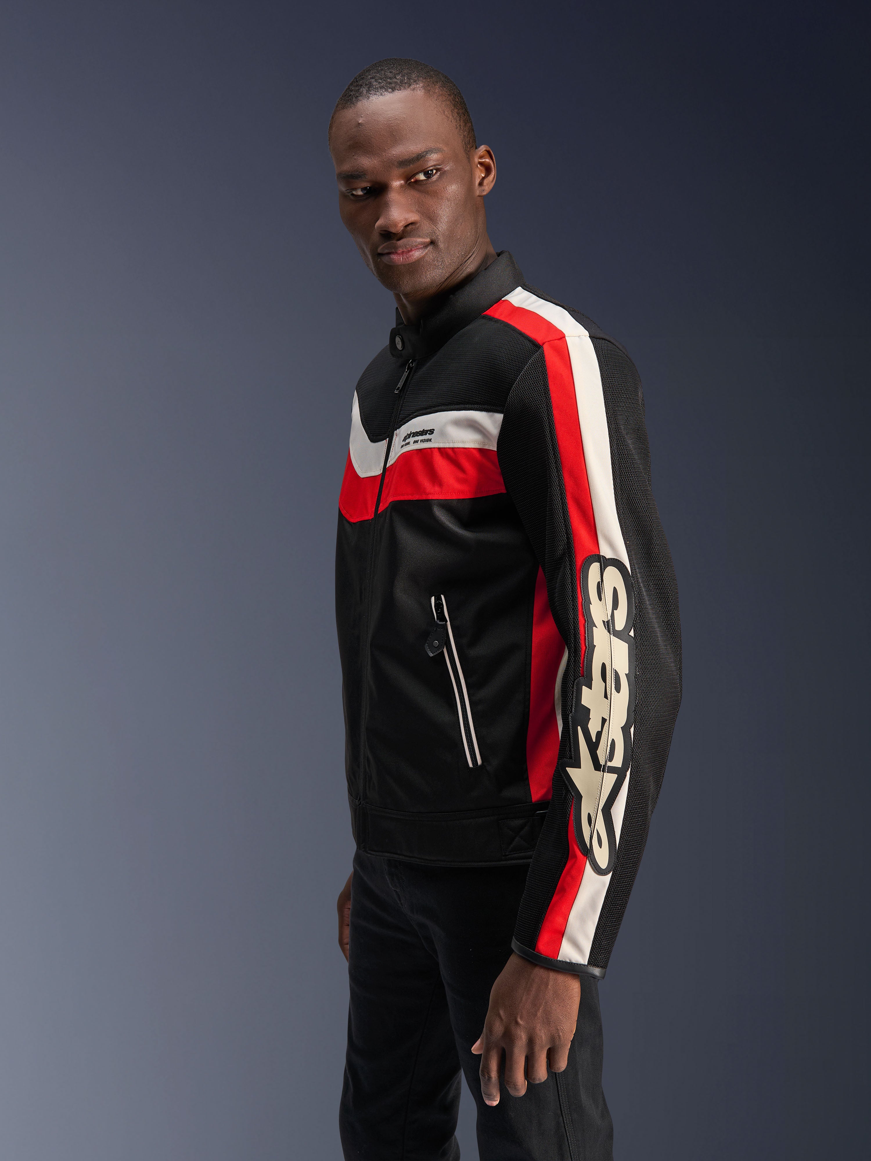 Modelo masculino con la chaqueta Alpinestars T-Dyno Air en negro, rojo brillante y crudo, chaqueta de moto textil de inspiración vintage con paneles de malla 3D, rayas llamativas en rojo y blanco hueso, y logotipo de Alpinestars destacado en la manga