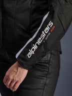 Chaqueta Alpinestars Stella T-SPS Air V2, chaqueta de moto para mujer, negra y blanca, primer plano de la manga con el logotipo blanco de Alpinestars y el texto T-SPS AIR, confección en malla y tejido técnico para favorecer el flujo de aire, usada con pantalones de moto negros