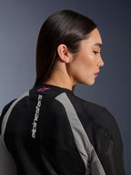 Perfil en primer plano de una mujer vistiendo la chaqueta Alpinestars Stella Troop-Air, confección de malla en negro y gris ceniza, logotipo vertical blanco de Alpinestars en la parte superior de la espalda con logotipo de estrella en rosa flúor, equipamiento técnico ligero de motociclismo para mujer