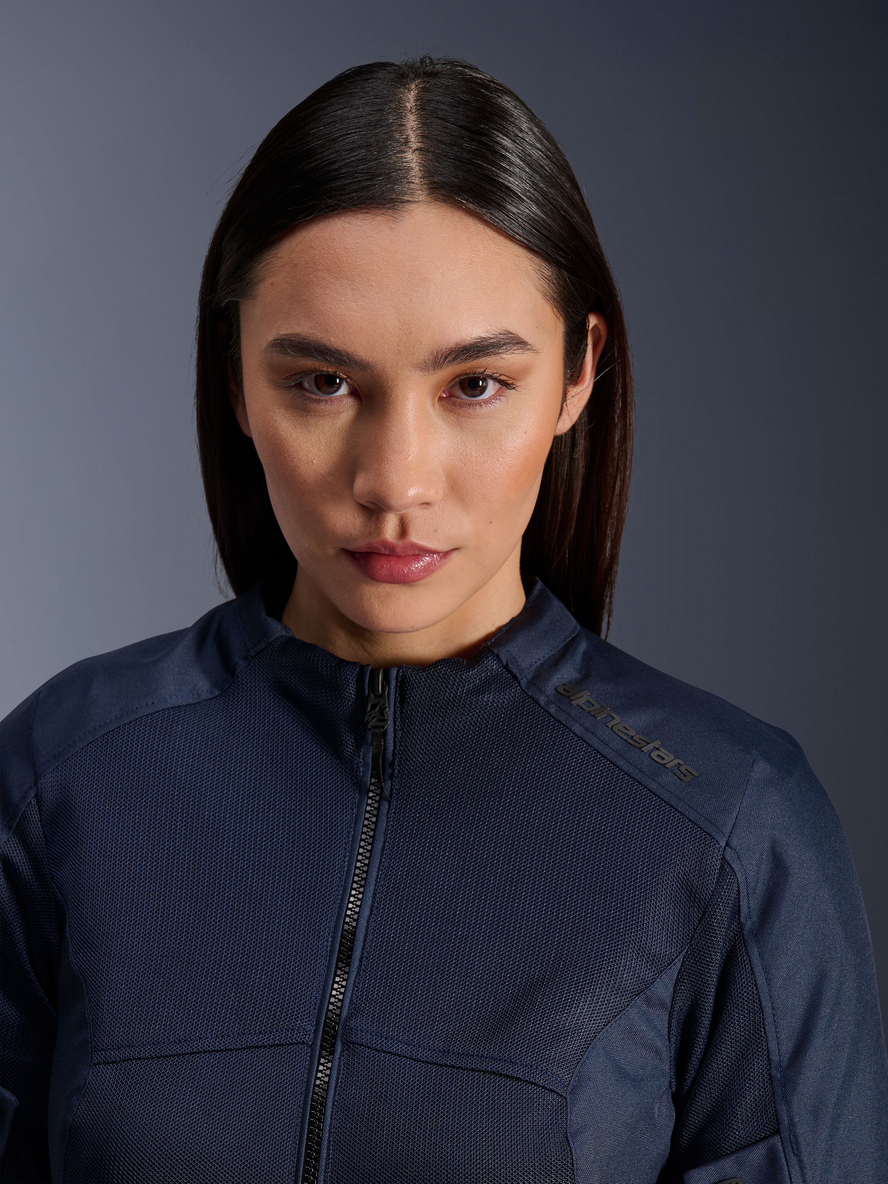 Modelo femenina luciendo la chaqueta Stella C-1 Air de Alpinestars en azul oscuro, primer plano que muestra la textura de tejido de malla transpirable, cremallera frontal y logotipo de Alpinestars en el cuello, elegante prenda de motociclismo urbano