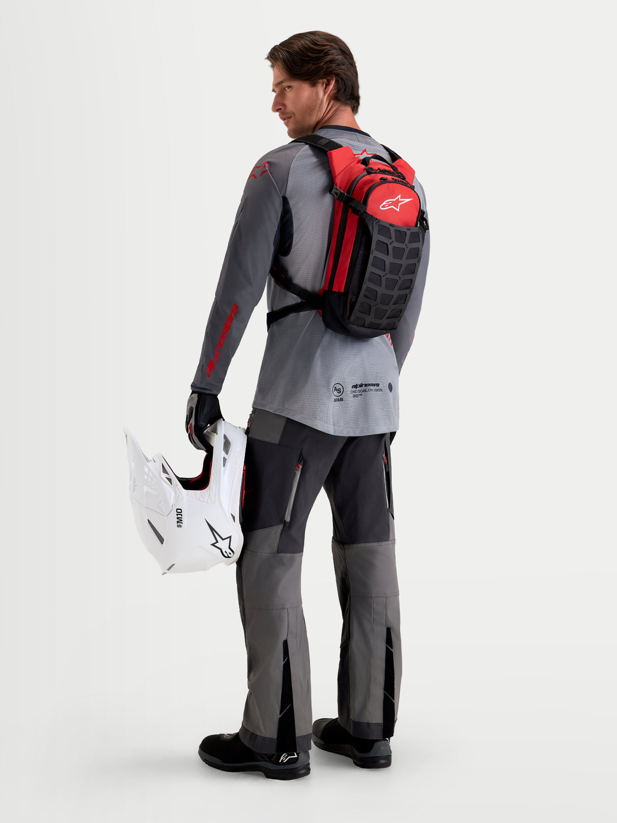 Alpinestars AT-9 Backpack, motocross backpack in black and bright red, worn by a model from the rear holding a white MX helmet and matching gear, lightweight textile build for track and paddock use