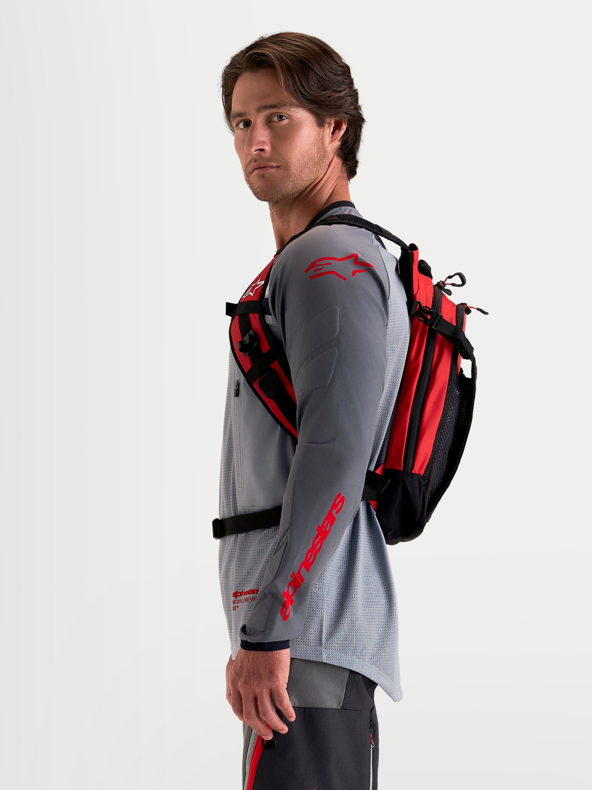 Alpinestars AT-9 Backpack, motocross backpack in black and bright red, side profile view worn by a male model wearing matching MX riding gear, featuring a lightweight textile construction and ergonomic harness system