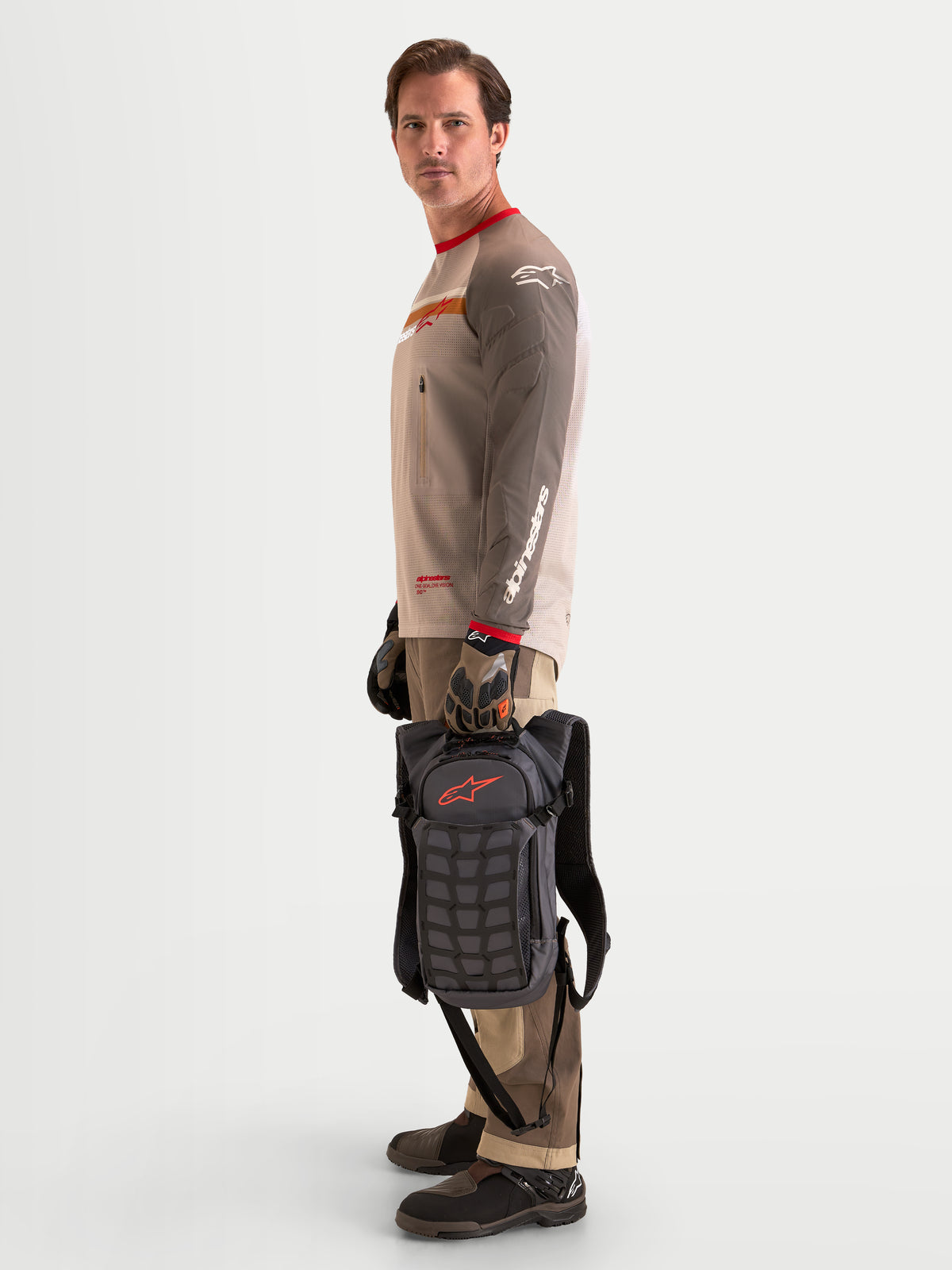 Alpinestars AT-9 Backpack, motocross backpack in walnut and deep gray, side profile view of a model standing and holding the lightweight textile gear pack by its top handle, showcasing structural paneling and organized storage compartments