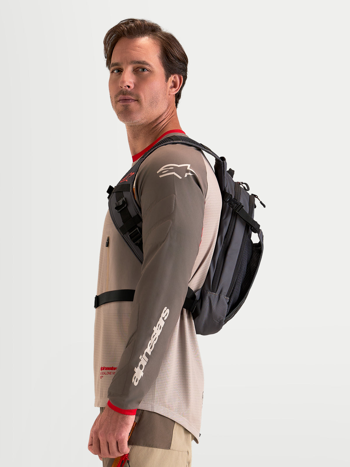 Alpinestars Mochila AT-9, mochila de motocross en color marrón y gris oscuro, vista lateral con modelo masculino mostrando el perfil del arnés y la construcción textil ligera