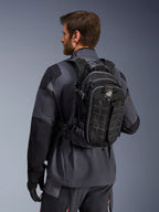 Mochila Alpinestars AT-15, mochila de aventura para motocicleta, negra y gris oscuro, llevada por un piloto con una chaqueta gris, con un diseño técnico con cinchas MOLLE y marca 'TERRA', equipamiento ergonómico para off-road
