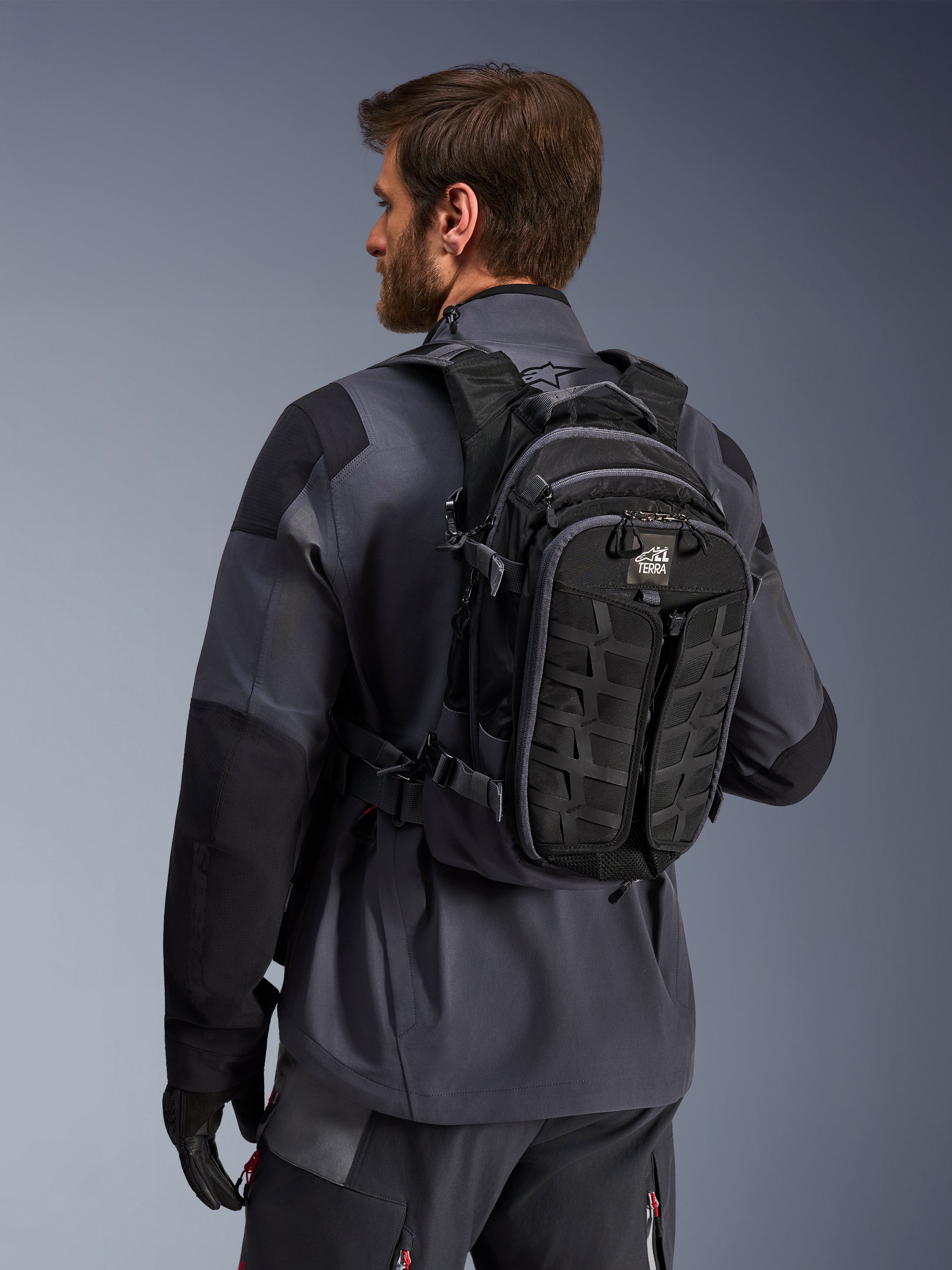Mochila Alpinestars AT-15, mochila de aventura para motocicleta, negra y gris oscuro, llevada por un piloto con una chaqueta gris, con un diseño técnico con cinchas MOLLE y marca 'TERRA', equipamiento ergonómico para off-road