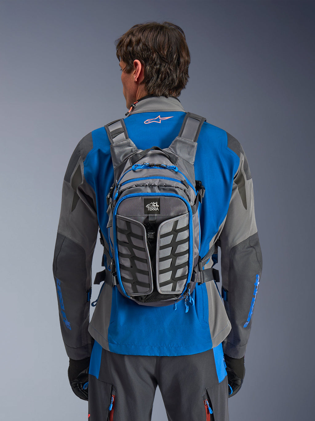 Mochila Alpinestars AT-15, mochila de motociclismo de aventura, gris oscuro y gris profundo, llevada por un piloto sobre una chaqueta azul y gris, con la marca All Terra, diseño estructural ergonómico, correas ajustables y detalles de cremalleras en azul.