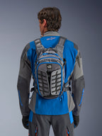 Mochila Alpinestars AT-15, mochila de motociclismo de aventura, gris oscuro y gris profundo, llevada por un piloto sobre una chaqueta azul y gris, con la marca All Terra, diseño estructural ergonómico, correas ajustables y detalles de cremalleras en azul.