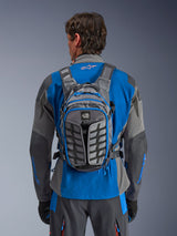 Mochila Alpinestars AT-15, mochila de motociclismo de aventura, gris oscuro y gris profundo, llevada por un piloto sobre una chaqueta azul y gris, con la marca All Terra, diseño estructural ergonómico, correas ajustables y detalles de cremalleras en azul.