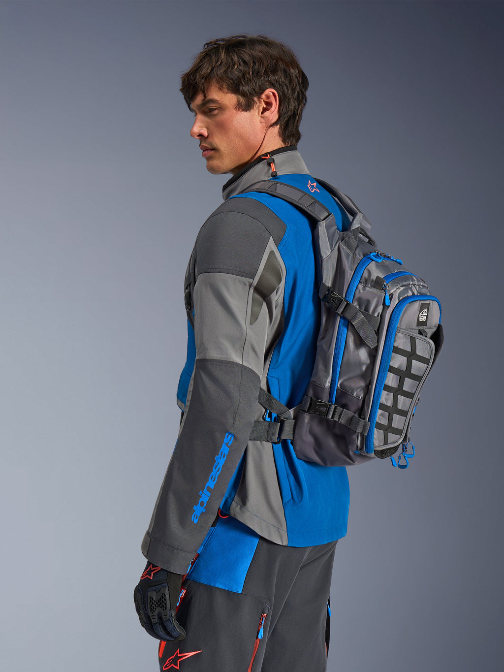 Mochila Alpinestars AT-15, mochila de aventura para moto, gris oscuro con detalles en azul, llevada por un piloto con equipación técnica coordinada en gris y azul, con panel MOLLE y construcción robusta para aventuras off-road.