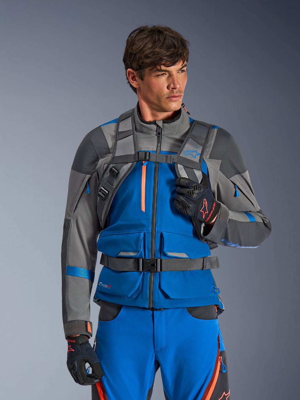 Alpinestars AT-15 Back Pack, mochila para moto, gris oscuro, llevada por un piloto con chaqueta y pantalones de aventura en azul y gris, con correas técnicas grises en los hombros, hebilla magnética en el pecho y correa de cintura, diseño robusto para conducción off-road
