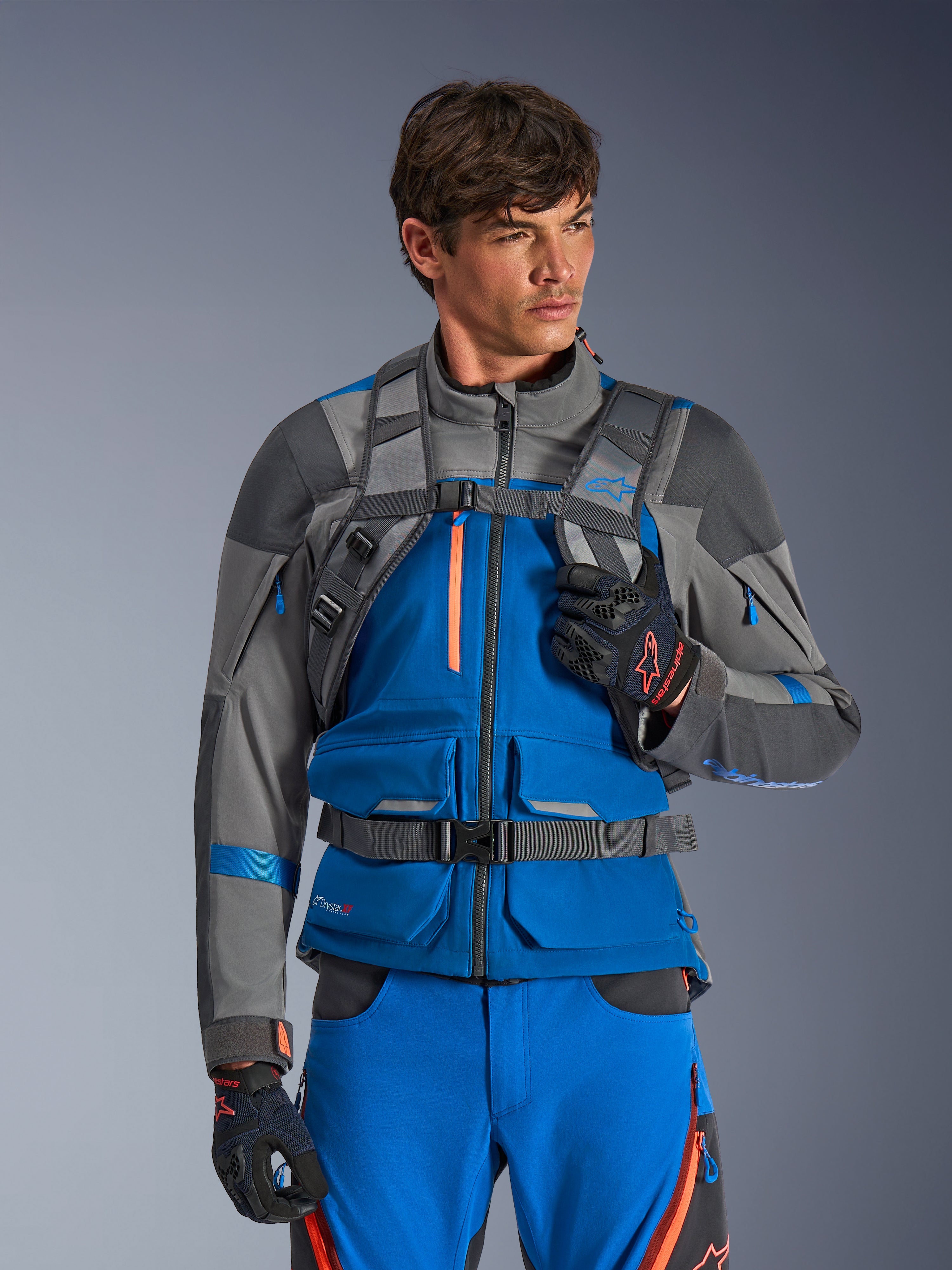 Alpinestars AT-15 Back Pack, mochila para moto, gris oscuro, llevada por un piloto con chaqueta y pantalones de aventura en azul y gris, con correas técnicas grises en los hombros, hebilla magnética en el pecho y correa de cintura, diseño robusto para conducción off-road