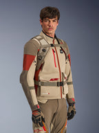 Mochila Alpinestars AT-15 en Aluminum Walnut, usada por un piloto sobre una chaqueta de moto adventure a juego, mostrando el arnés frontal con cierre magnético en el pecho y correas ajustables, equipamiento técnico en arena y rojo para conducción de aventura