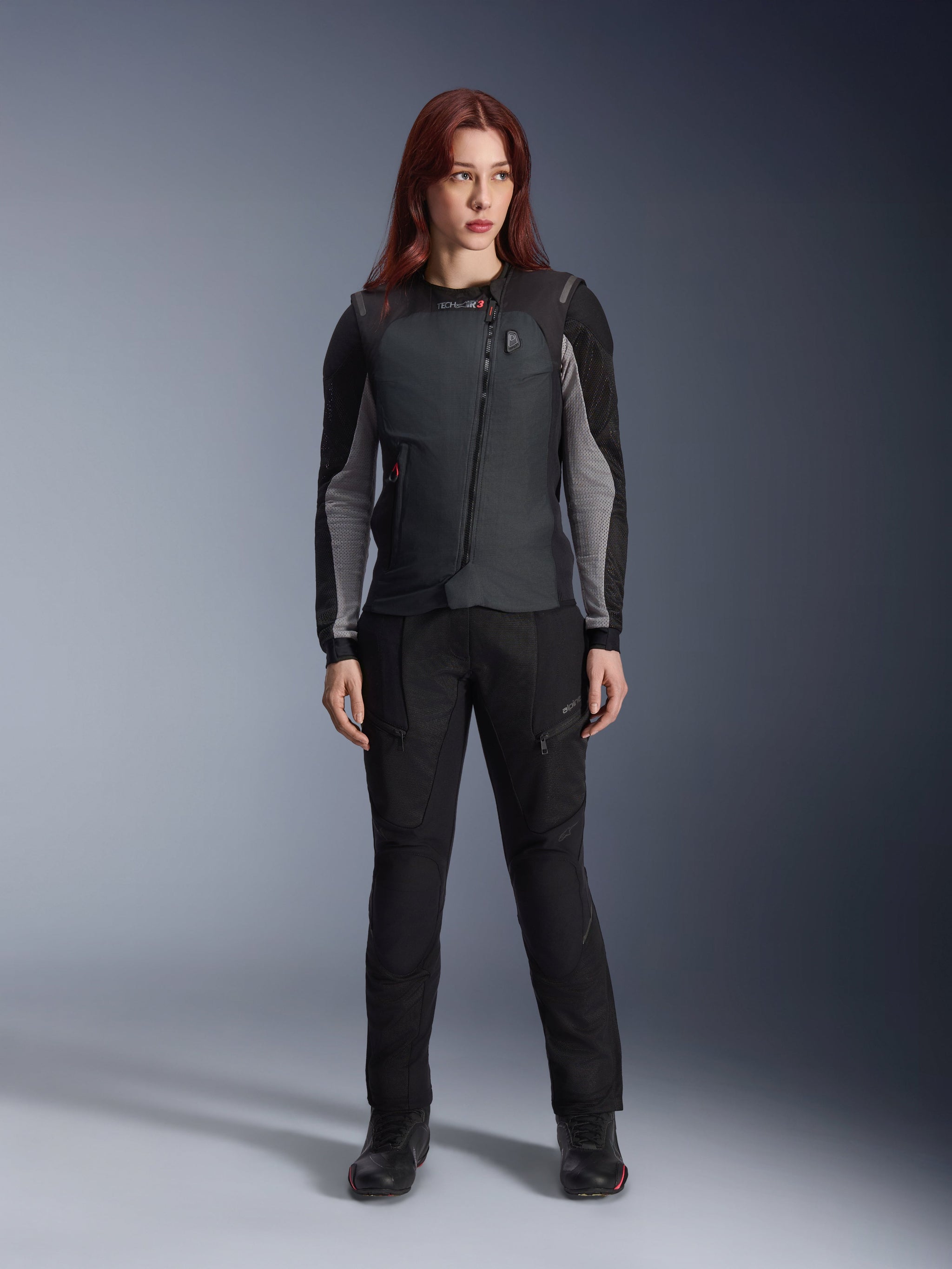 Sistema Alpinestars Stella Tech-Air® 3 V2 para mujer, chaleco airbag de moto negro, usado sobre una capa técnica de manga larga negra y gris con pantalones de conducción negros a juego, con cremallera diagonal y pantalla LED de estado, ajuste femenino entallado para motociclistas de carretera y turismo