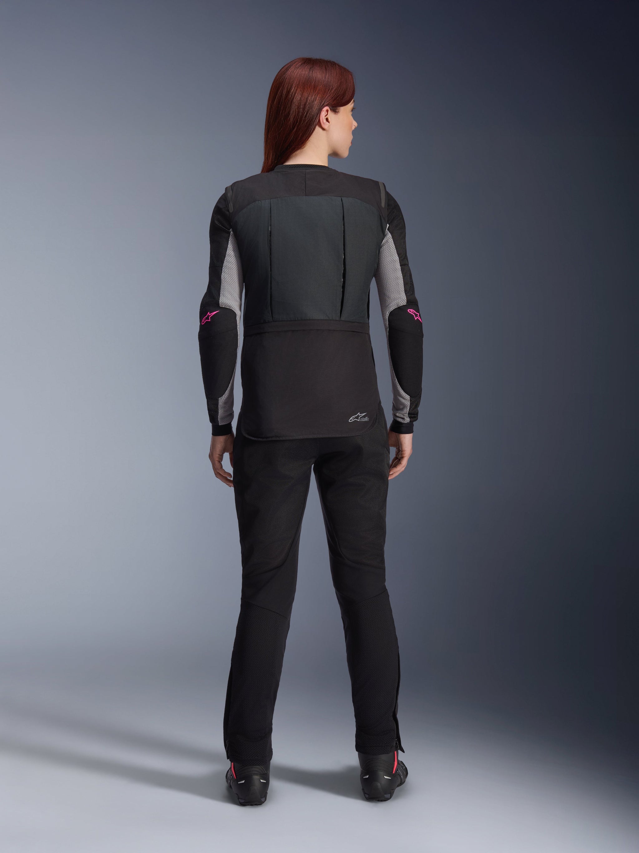 Vista trasera de una mujer con el sistema Alpinestars Stella Tech-Air® 3 V2 en color negro, chaleco airbag autónomo para moto, combinado con pantalones de conducción negros y capa base con logotipos rosas, mostrando el ajuste femenino estilizado y entallado para conducción urbana y en carretera.