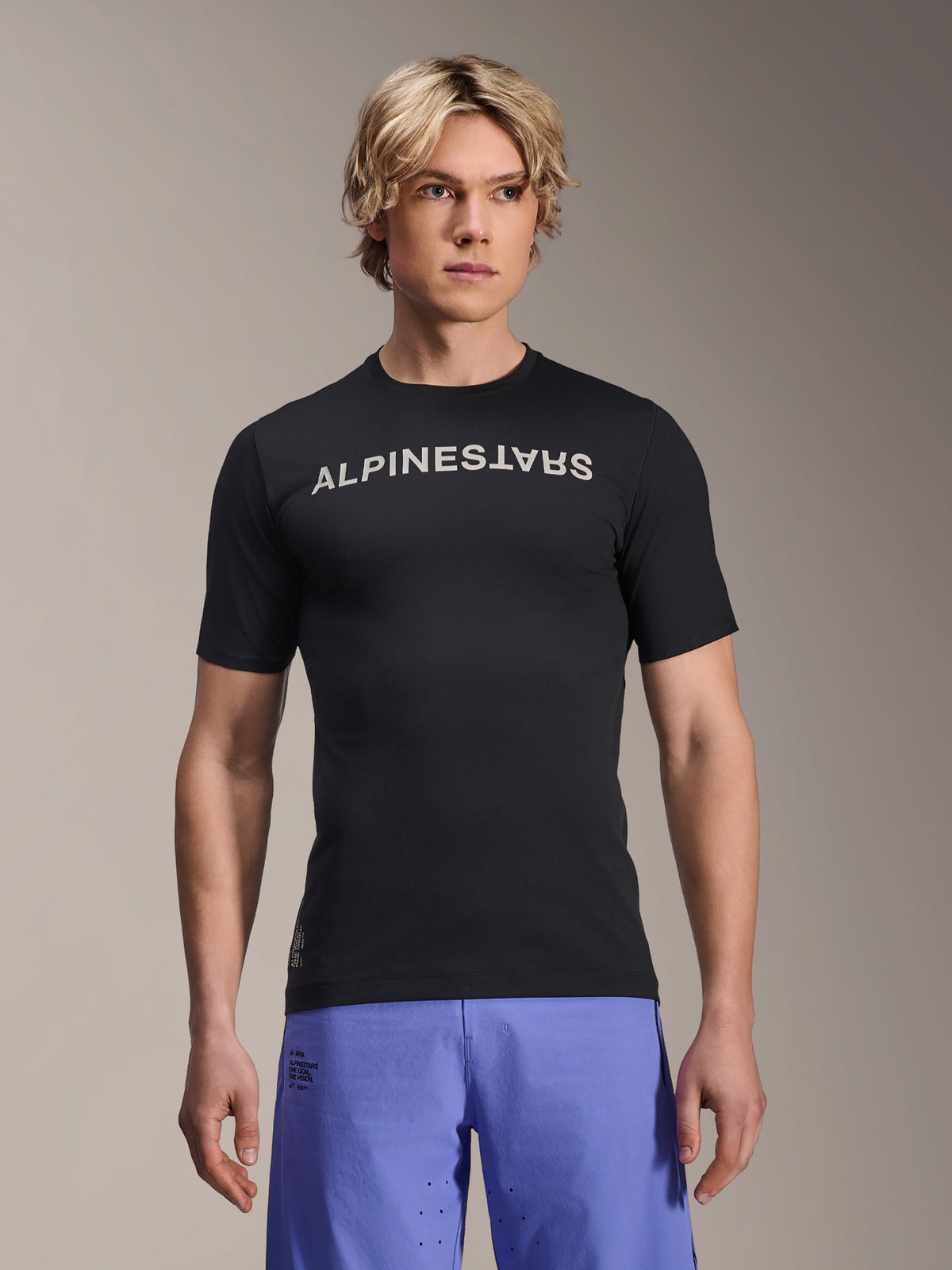 Alpinestars A-Aria Switch Jersey, camiseta de manga corta para MTB, negra con marca denominativa Alpinestars en blanco en el pecho, usada por un modelo masculino con pantalones cortos morados, tejido ligero de alto rendimiento para trail