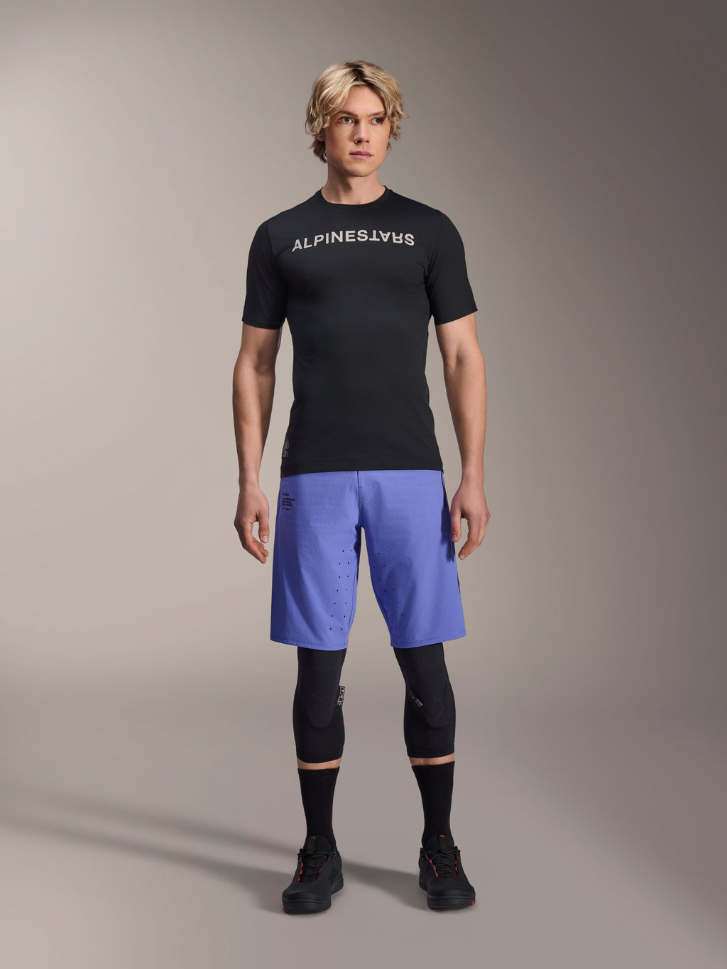 Alpinestars A-Aria Switch Jersey, Short Sleeve MTB Jersey, Black with mirrored white Alpinestars logo, worn by a male model with blue cycling shorts and black knee pads, breathable performance apparel for trail riding