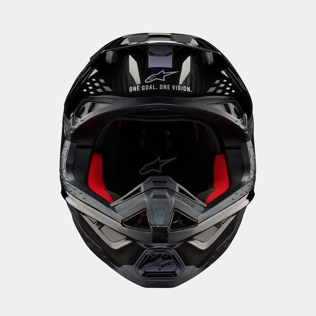 Casco Alpinestars Supertech M10 Solid, casco de motocross, carbono negro brillante, vista frontal que muestra el forro interior rojo, calota de fibra de carbono y el eslogan «ONE GOAL. ONE VISION.» en el área de la visera