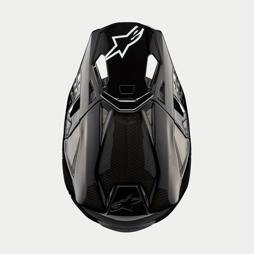 Casco Alpinestars Supertech M10 Solid, casco de motocross, carbono negro brillante, vista superior que muestra el tejido de fibra de carbono visible, logotipo blanco de Astars en la visera, perfil aerodinámico estilizado con puertos de ventilación integrados