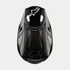 Casco Alpinestars Supertech M10 Solid, casco de motocross, carbono negro brillante, vista superior que muestra el tejido de fibra de carbono visible, logotipo blanco de Astars en la visera, perfil aerodinámico estilizado con puertos de ventilación integrados