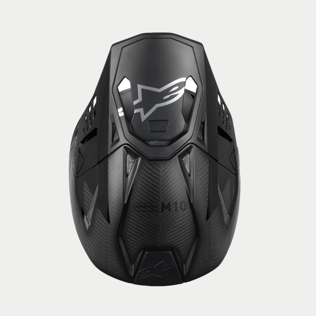 Casco Alpinestars Supertech M10 Fame ECE 22.06, casco de motocross, carbono negro mate y brillante, vista superior que resalta el tejido de fibra de carbono, visera aerodinámica con logotipo plateado de Alpinestars y múltiples puertos de ventilación para carreras todoterreno.