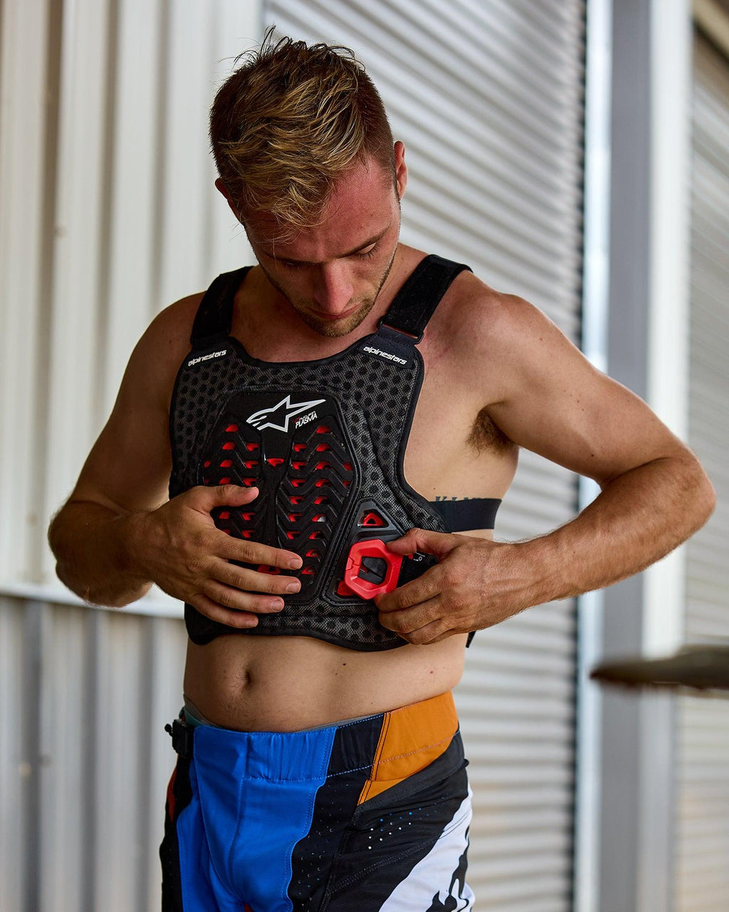 Hombre poniéndose el protector de pecho Alpinestars A-5 Plasma, negro con detalles rojos y logotipo blanco, con una estructura de malla de panal, usado con pantalones de motocross azules y naranjas, en un entorno de preparación para carreras todoterreno