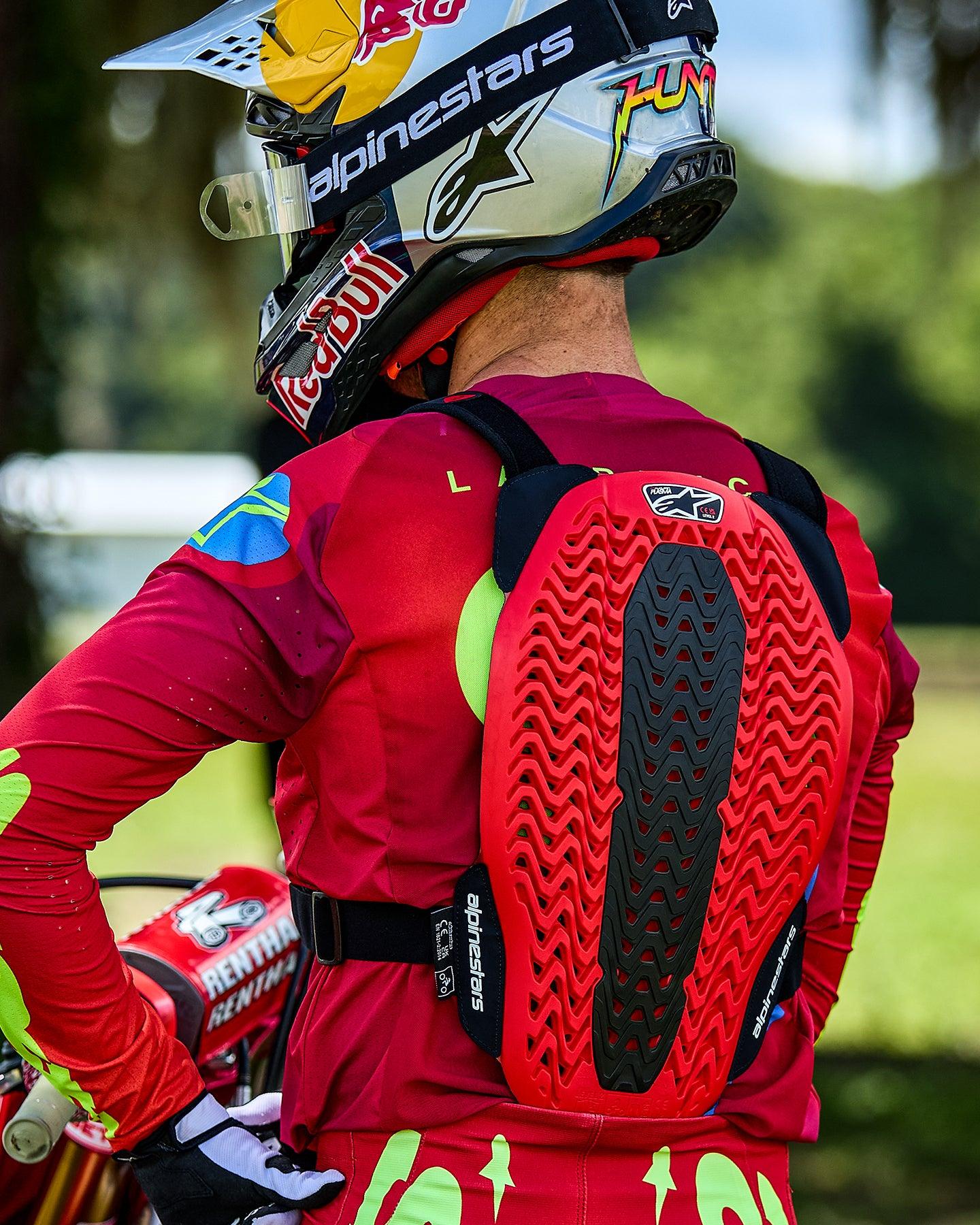 Alpinestars A-5 Plasma Chest Protector, protector de espalda para motocross visto desde atrás, con una estructura de panal ventilada en rojo y negro, usado sobre un jersey rojo de manga larga y un casco de la marca Red Bull.