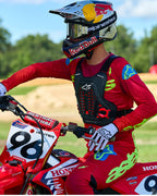 Piloto de motocross con el protector de pecho Alpinestars A-5 Plasma en negro y rojo, combinado con jersey y pantalones de carreras en rojo y verde lima, sentado sobre una motocicleta Honda roja, equipo de protección off-road de alto rendimiento