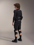Modelo vistiendo los protectores de rodilla y espinilla Alpinestars A-Impact Plasma Pro desde atrás, en negro y blanco, mostrando el cierre de triple correa y los paneles de malla ventilada, combinados con un equipo completo de protección para bicicleta de montaña en color negro que incluye capa base de manga larga y pantalones cortos acolchados.