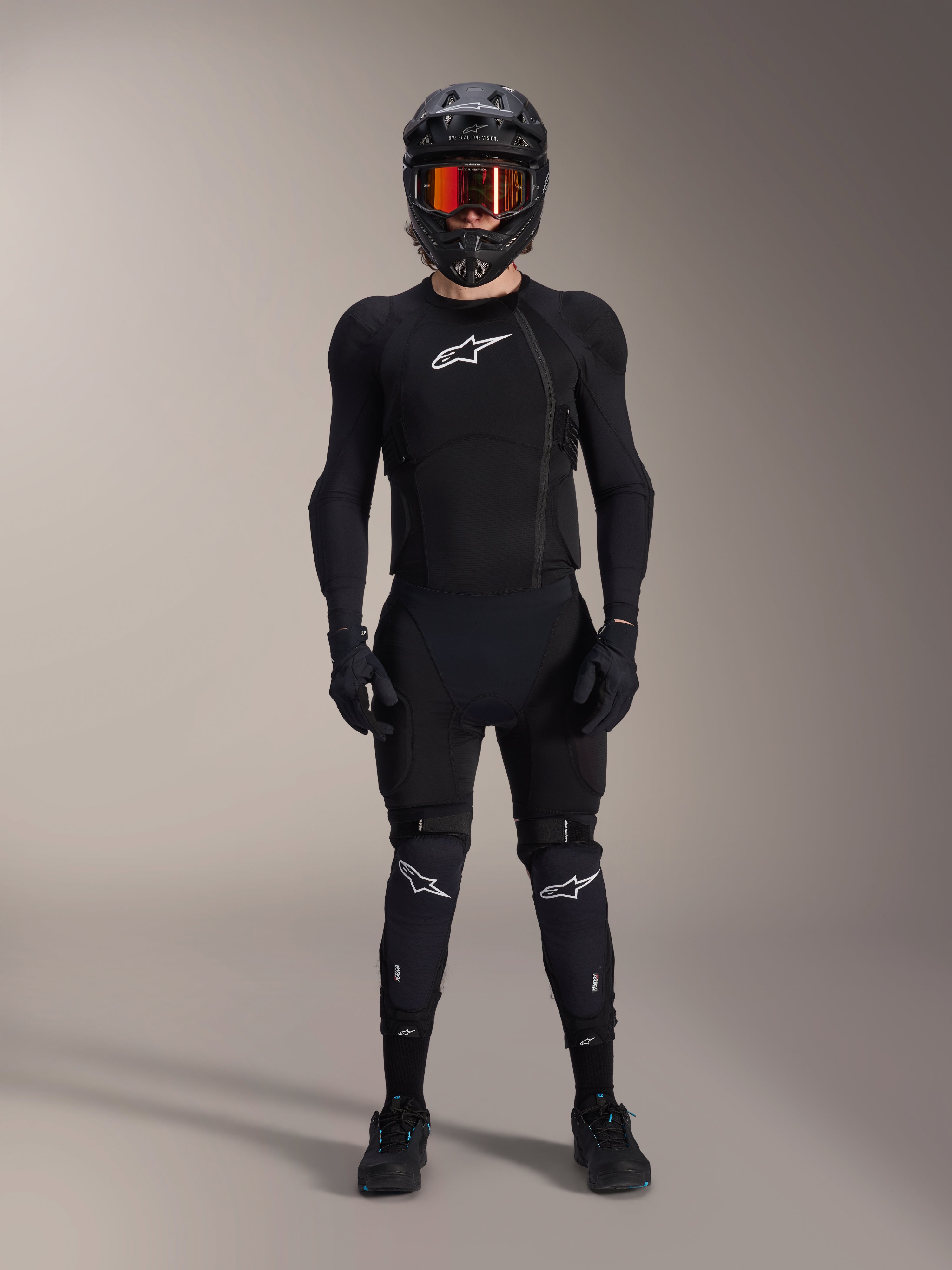 Ciclista de montaña con equipamiento de protección integral, chaqueta de protección Alpinestars A-Impact Plasma en negro y blanco, con logotipo blanco en el pecho y tejido de malla, acompañada de casco negro, gafas de lentes rojas y rodilleras, conjunto de armadura de MTB coordinado de alto rendimiento.