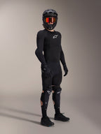 Protectores de rodilla y espinilla Alpinestars A-Impact Plasma Pro, en negro y blanco, usados por un ciclista con un equipamiento protector completo para bicicleta de montaña, que incluye una prenda superior blindada de manga larga negra, guantes negros y un casco integral con gafas naranjas.