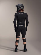 Protectores de rodilla y espinilla Alpinestars A-Impact Plasma Pro, negro, vista trasera de un ciclista con equipo de protección integral de MTB, incluyendo casco integral y armadura corporal, con sistema de cierre de triple correa y paneles de malla ventilada para un ajuste seguro y flujo de aire