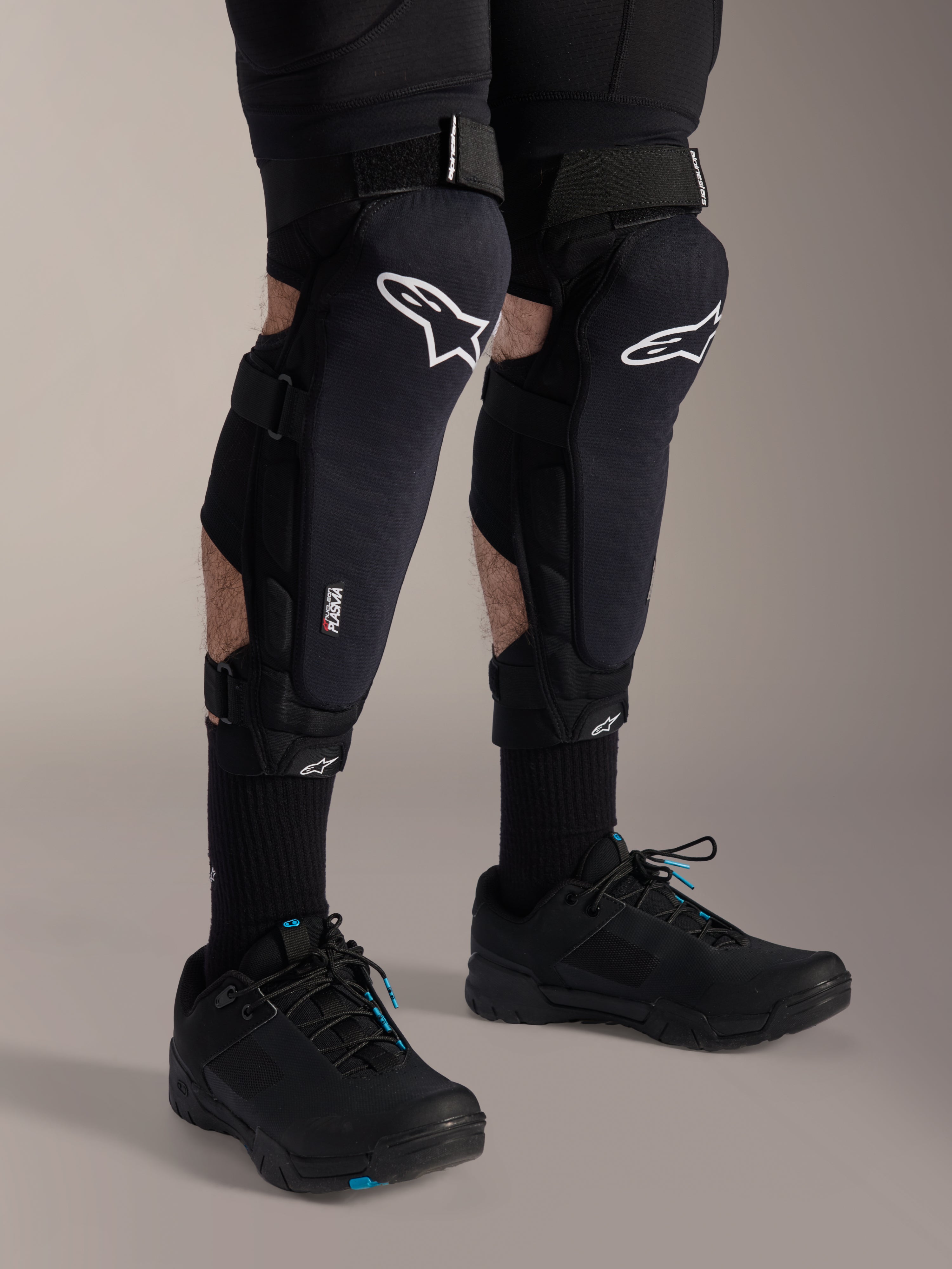 Protectores de rodilla y espinilla Alpinestars A-Impact Plasma Pro, negro y blanco, con el logotipo blanco de Alpinestars y sistema de cierre de triple correa, usados con calcetines negros y zapatillas deportivas negras, protección duradera para ciclismo todoterreno
