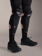 Rodilleras Alpinestars A-Impact Plasma Pro, negro y blanco, con logotipos de la tecnología Nucleon PLASMA, combinadas con calcetines negros y zapatillas de ciclismo de montaña negras, correas seguras de gancho y bucle, equipo de protección ergonómico para ciclismo de montaña
