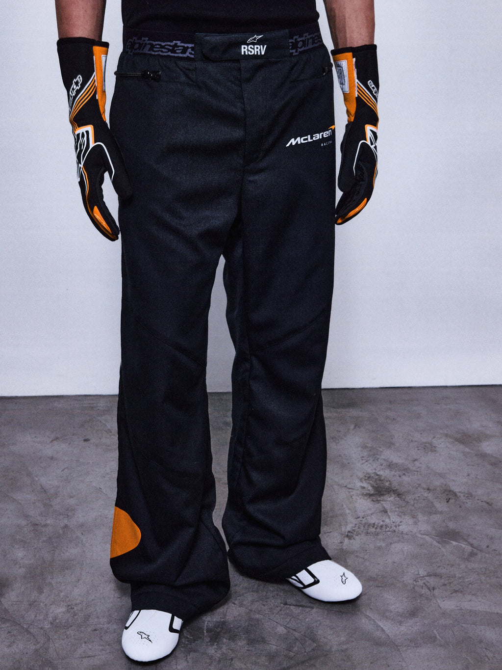 Alpinestars RSRV Arena Suit Pants, pantalones de carreras negros con parches naranjas, logos de McLaren y Alpinestars, persona usando guantes negros y naranjas, zapatos de carreras blancos con detalles negros
