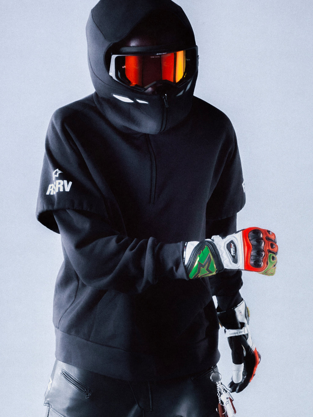 Alpinestars RSRV Vault Hoodie, sudadera negra con capucha, persona con casco y gafas reflectantes naranja-rojo, guantes de colores rojo, verde y blanco, logos blancos en la manga, estilo urbano futurista