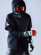 Alpinestars RSRV Vault Hoodie, sudadera negra con capucha, persona con casco y gafas reflectantes naranja-rojo, guantes de colores rojo, verde y blanco, logos blancos en la manga, estilo urbano futurista