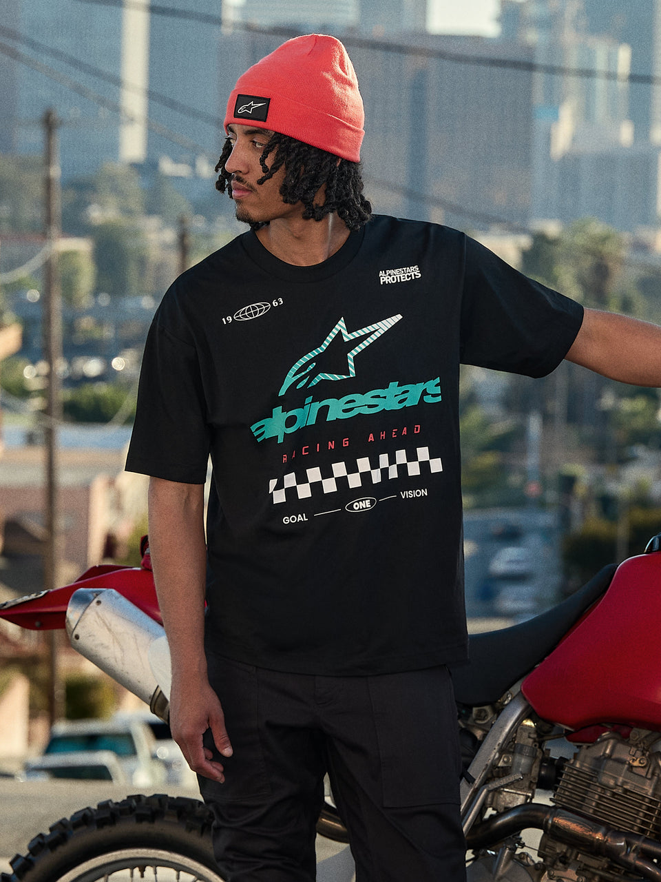 Alpinestars Aplomb Oversized CSF Tee, camiseta de manga corta, negra, con un gran logotipo Astars con degradado en verde azulado y blanco y un gráfico de bandera a cuadros, combinada con un gorro color coral, de pie junto a una motocicleta roja, estilo urbano casual