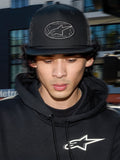 Gorra Alpinestars Contender Trucker, gorra negra de estilo de vida con parte posterior de malla de poliéster, corona estructurada con un parche bordado del logo Astars, llevada por un hombre con una sudadera con capucha negra de Alpinestars, ropa urbana casual inspirada en el automovilismo.