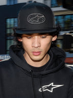 Gorra Alpinestars Contender Trucker, gorra negra de estilo de vida con parte posterior de malla de poliéster, corona estructurada con un parche bordado del logo Astars, llevada por un hombre con una sudadera con capucha negra de Alpinestars, ropa urbana casual inspirada en el automovilismo.